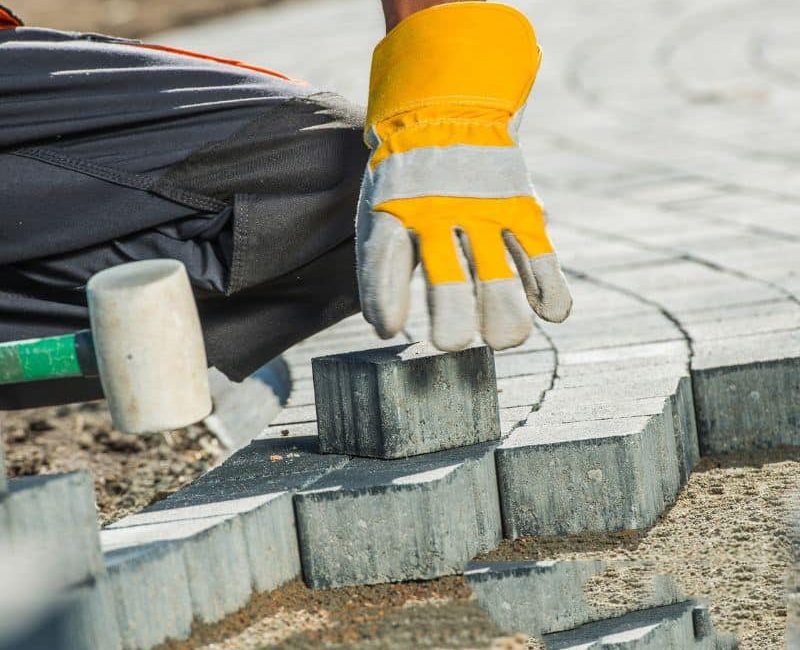 A man's hand in yellow gloves, laying bricks with a hammer for paving services in Sydney.