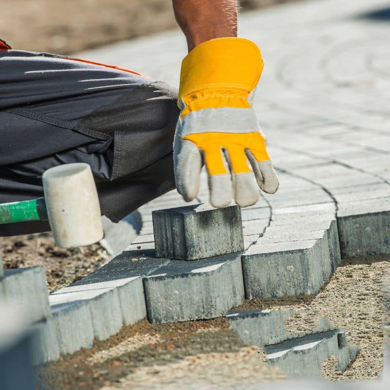A man's hand in yellow gloves, laying bricks with a hammer for paving services in Sydney.